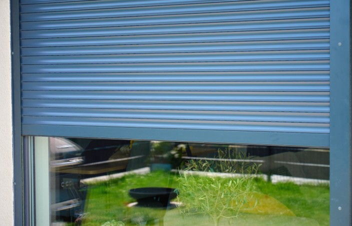 Persiana de aluminio a medida bajada a media altura sobre un ventanal, con el jardín reflejado en el cristal